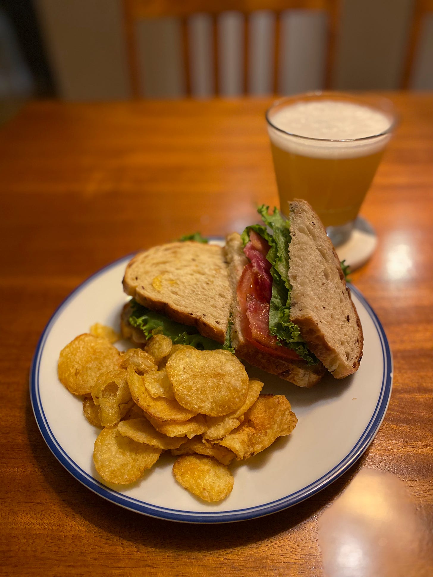 A white plate with a blue rim holds a sandwich on grainy sourdough, sliced in half. One open half faces upright and inside is bacon, lettuce, and tomato. In front of it is a pile of potato chips, and in the background on a coaster is a glass of beer.