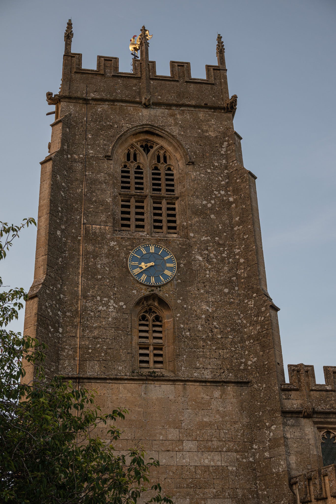 St Peter’s Church tower in Winchcombe, England with the blue-faced clock at 8:39 PM against a blue darkening evening sky