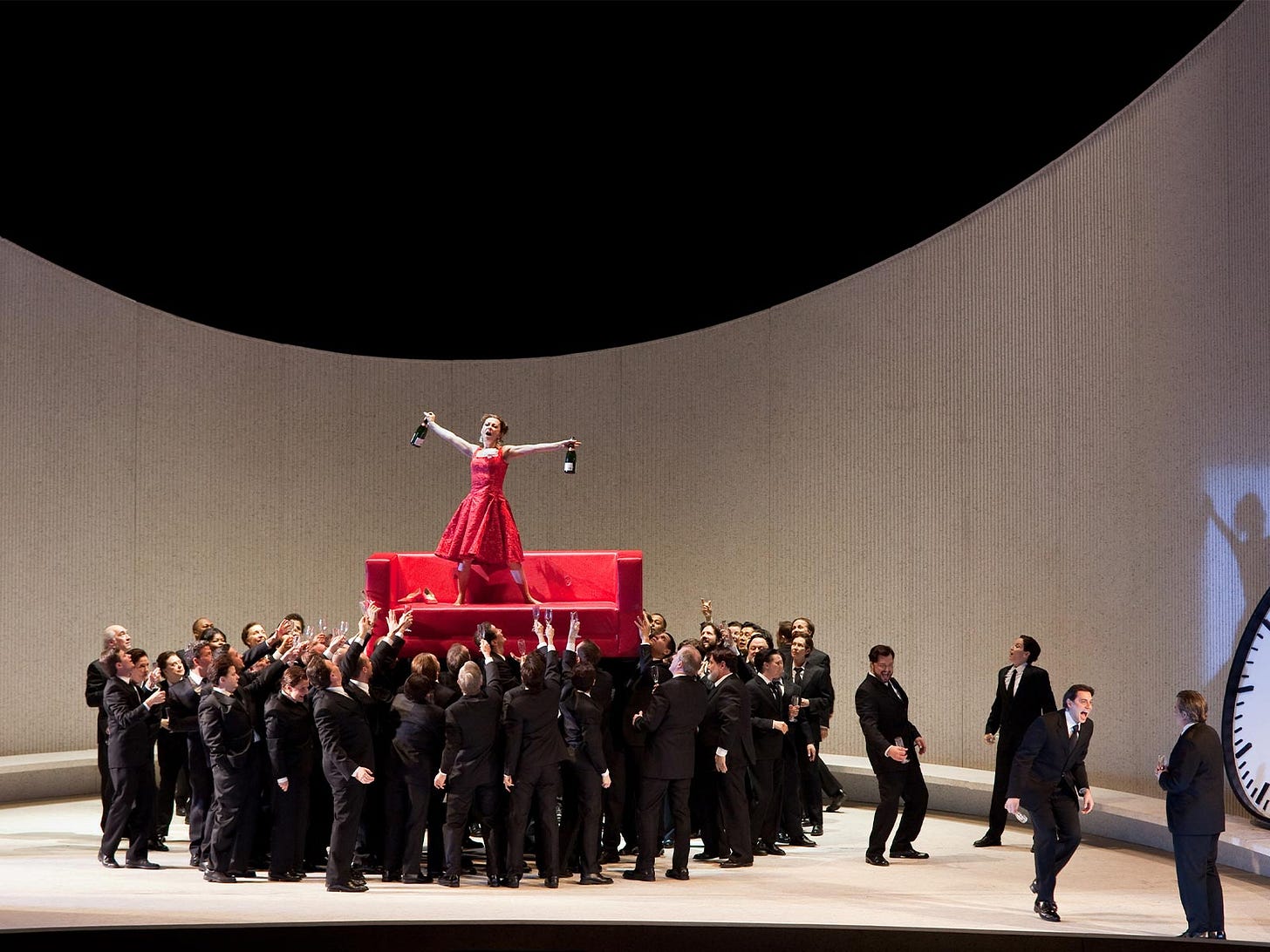 Violetta Valery, dressed in a hot pink party dress and standing on a matching couch, clasps a bottle of wine in each outstretched hand. A large chorus of men in tuxedos hoists her into the air. The stage setting is a sharply curved white wall, with a pitch black ceiling. Violetta Valery, dressed in a hot pink party dress and standing on a matching couch, clasps a bottle of wine in each outstretched hand. A large chorus of men in tuxedos hoists her into the air. The stage setting is a sharply curved white wall, with a pitch black ceiling.