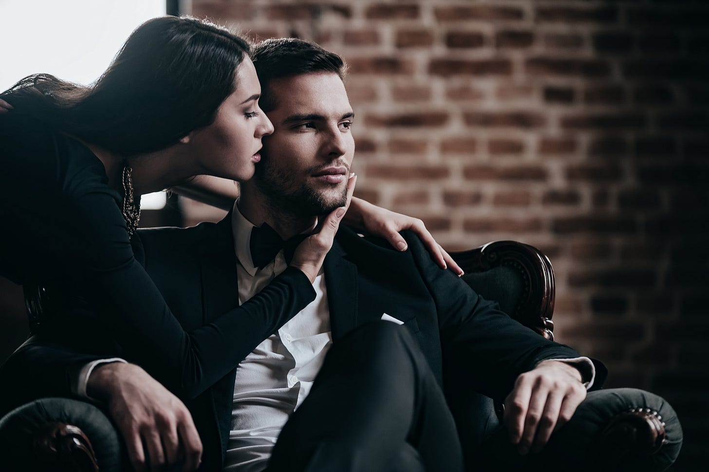 A woman dressed in black leans in closely and whispers in a man’s ear.