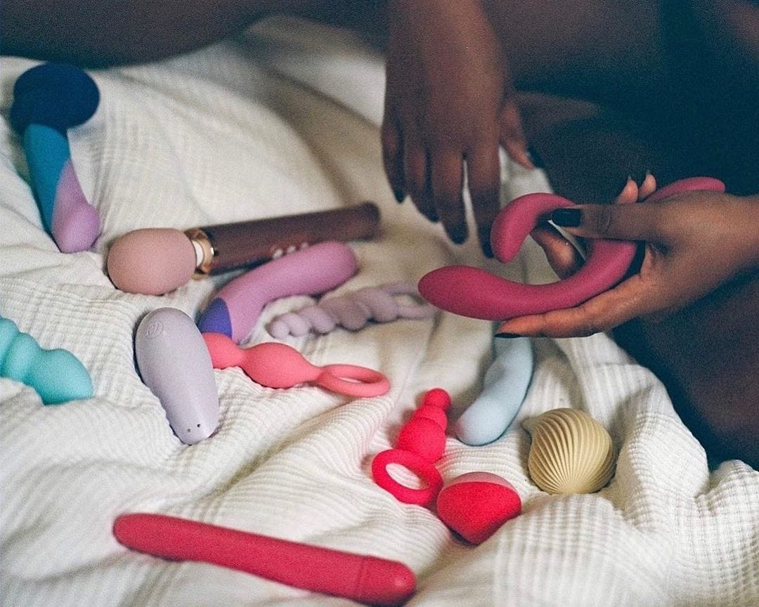 A photo of 12 pleasure products (vibrators, dildos, etc) spread out on a bed with hands touching them.
