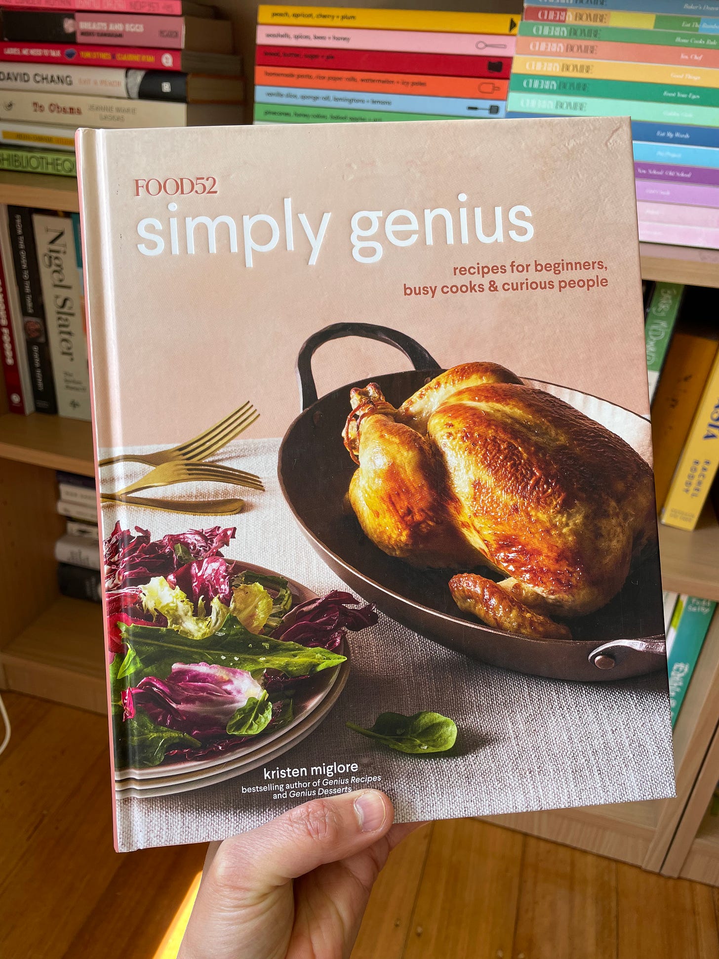 Food52 Simply Genius cookbook in front of a bookshelf filled with food magazines and books.