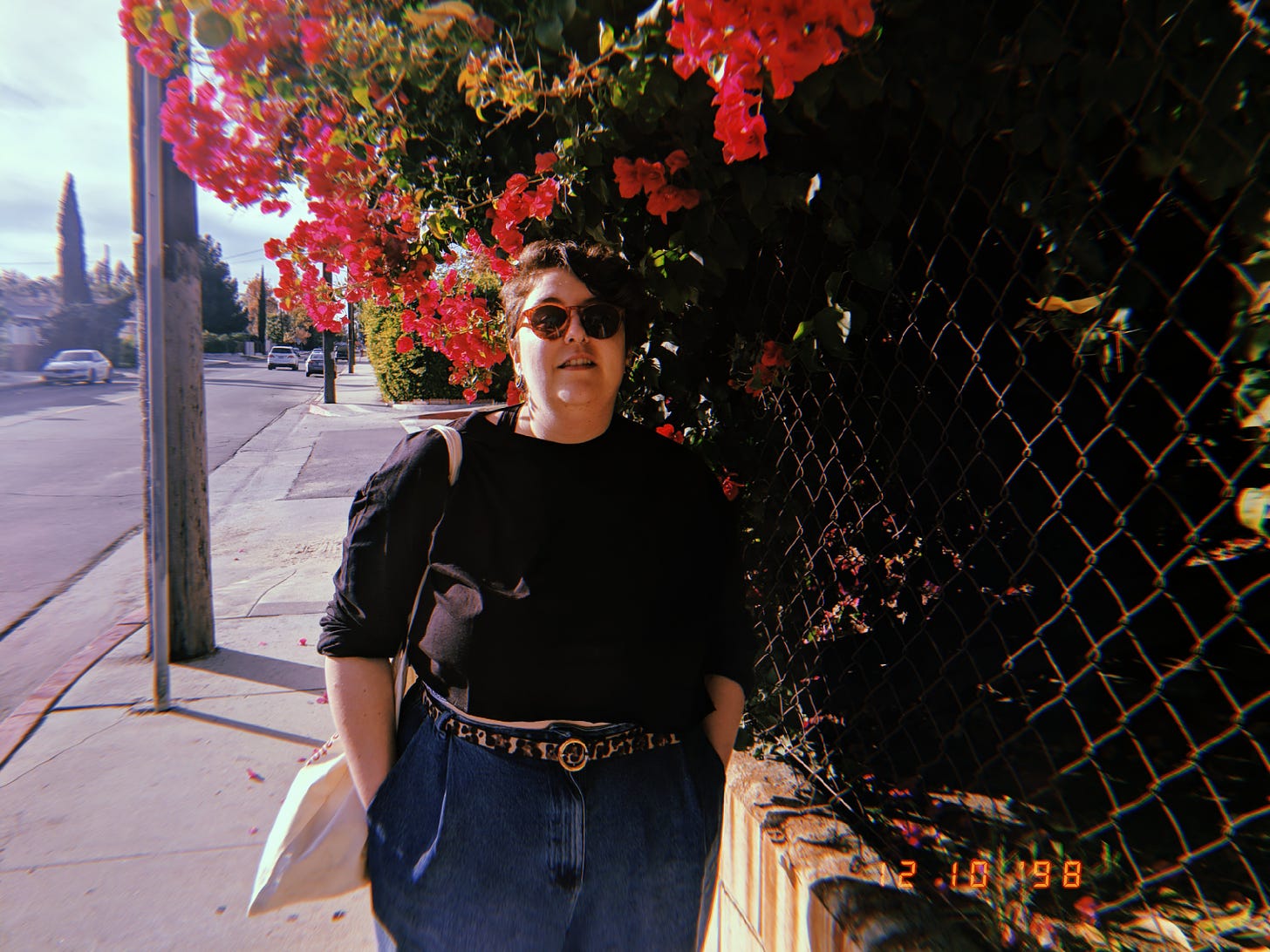 An image of me, a white non-binary person standing outside. I have on a black, long sleeve shirt, a leopard print belt, and dark high waisted jeans. My hands are in my pockets and there's a tote bag draped over my shoulder. I also have on sunglasses that are brown. I am smiling slightly, and have short brown hair that's longer on the right side. Next to me is a wire fence with a pink floral bush poking through.