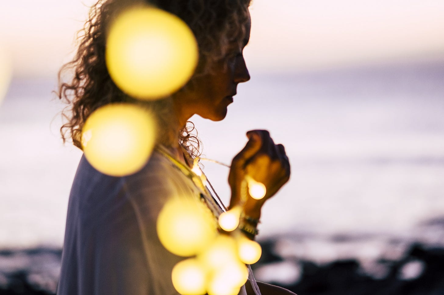 A woman, who is just out of focus, clutches a string of lights on a twilight beach.