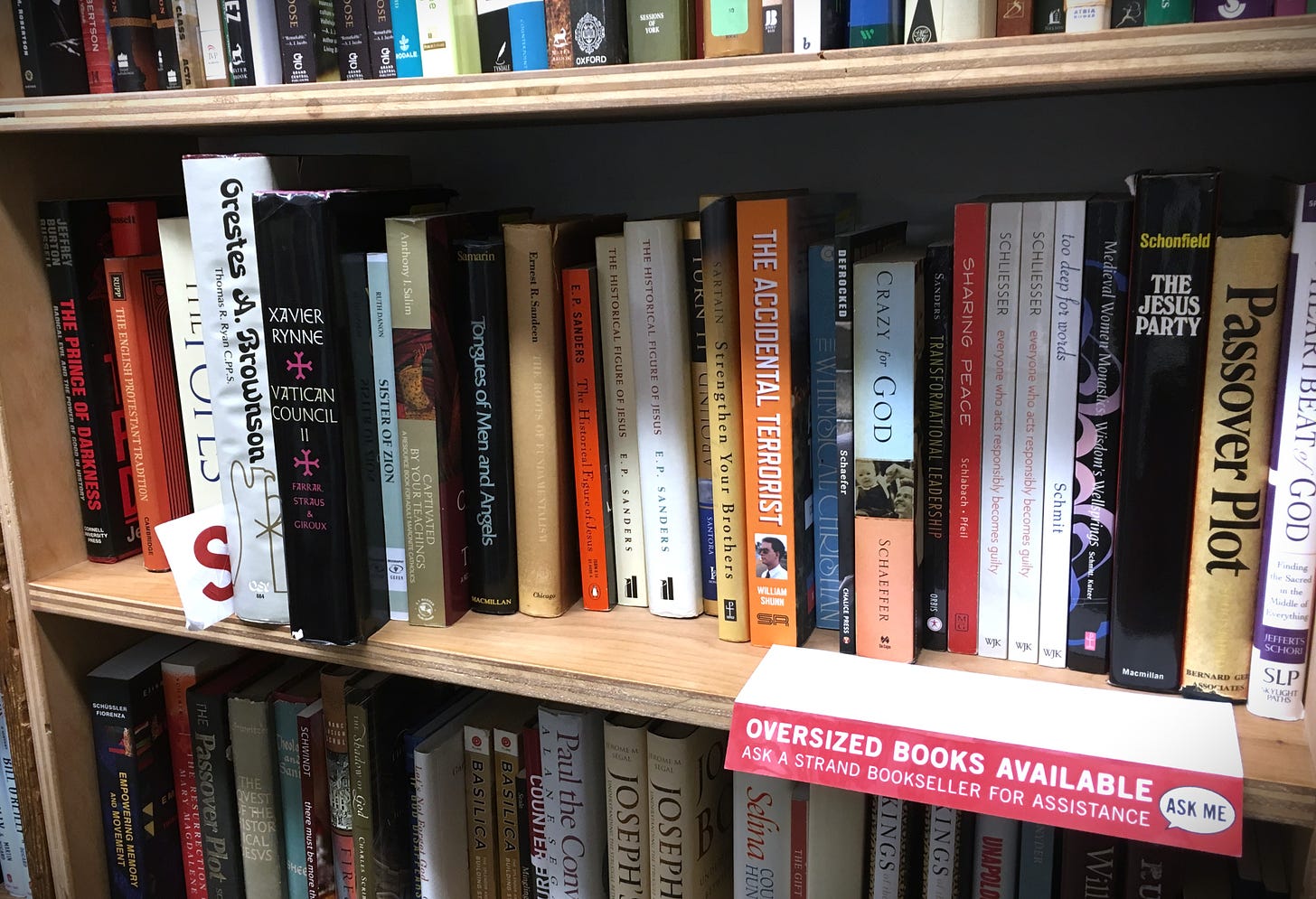 The spine of William Shunn's memoir, The Accidental Terrorist, sits on a crowded bookstore shelf amongst other religion-themed books. A sign on the shelf reads "Oversized Books Available. Ask a Strand bookseller for assistance."