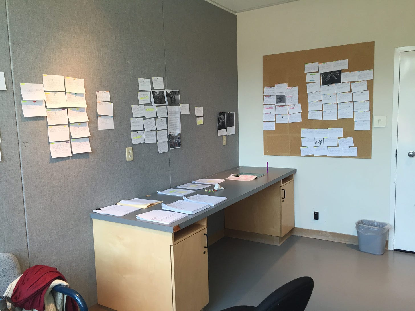 Photo of two walls of a room and a desk in front; the walls have index cards pinned all over them (one wall is fabric and the other has a bulletin board), and the desk is covered in tidy stacks of papers