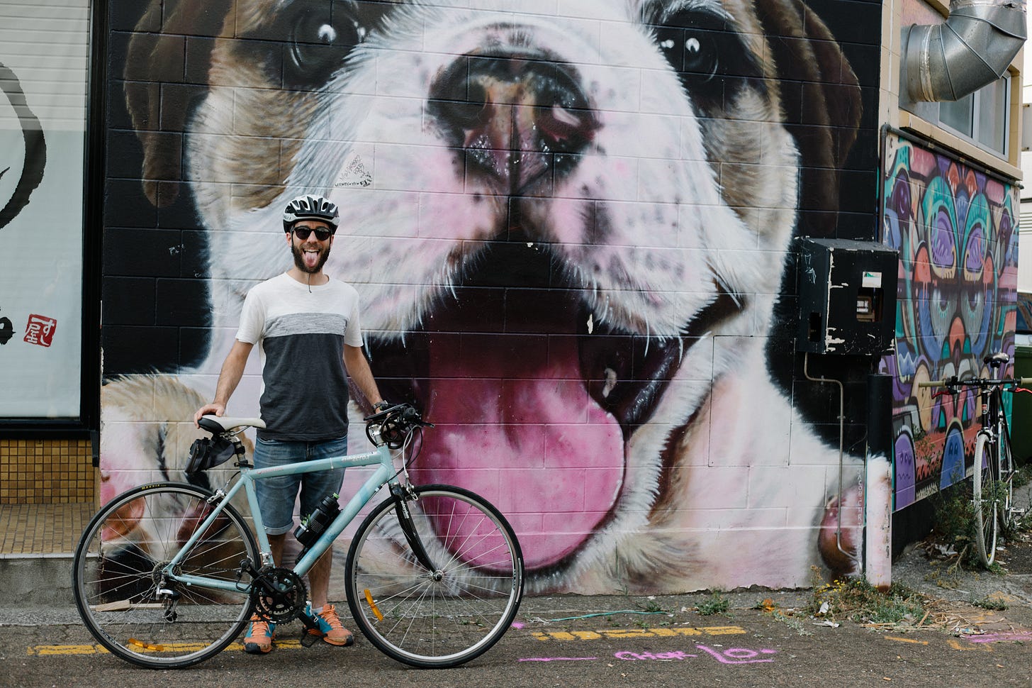 A photograph of a person standing with their bike in front of a large mural of a puppy with their tongue out. The person is standing in front, with their expression mirroring the puppy's in the mural. 