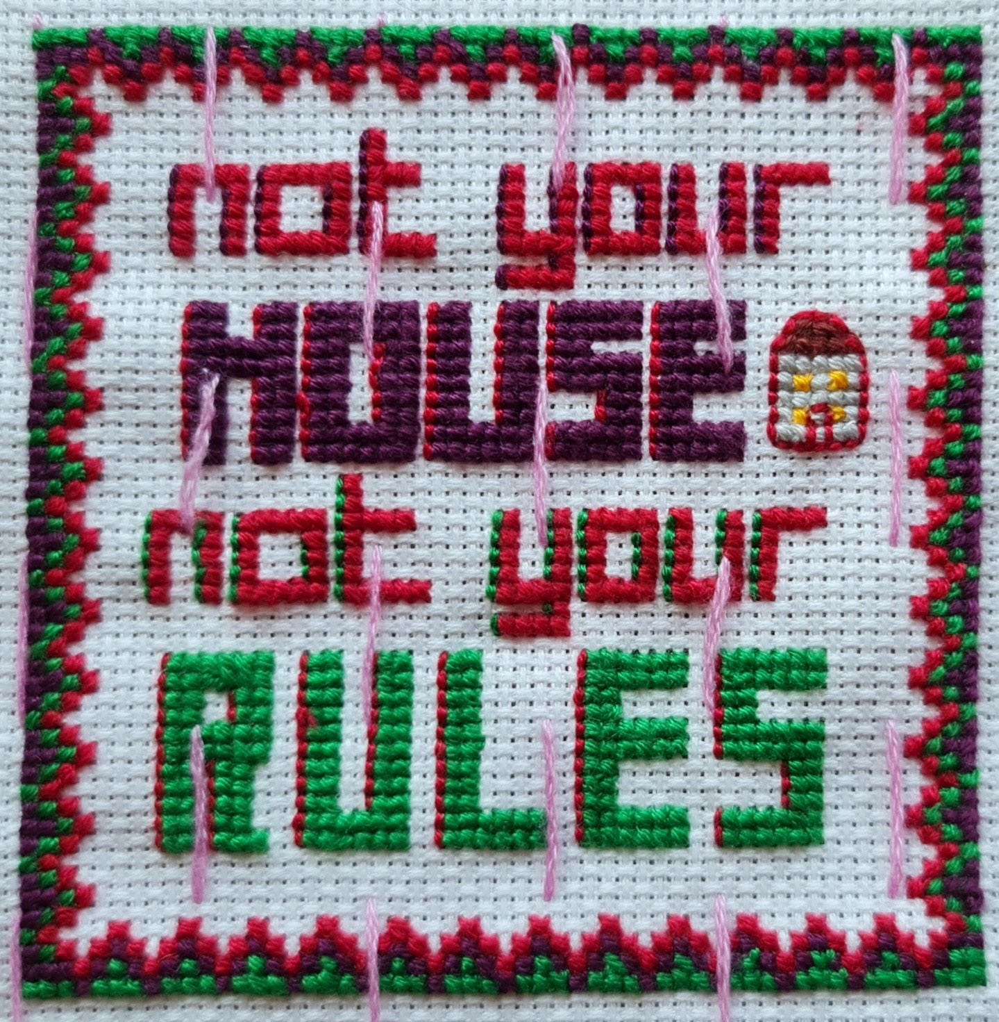 image description: a photograph of a square piece of white cross-stitch fabric with stitches spelling out “not your house not your rules”, a patterned border, and a tiny icon of a house. ‘not your’ is stitched in red with purple highlight edges along the left side of the letters. ‘house’ is stitched in purple with red highlight edges along the left. ‘rules’ is stitched in green with red highlight edges. the square border pattern has a repeated, stepped design, with the left/right edges starting in purple and going through green and red, and the top/bottom starting in green and going through purple and red.