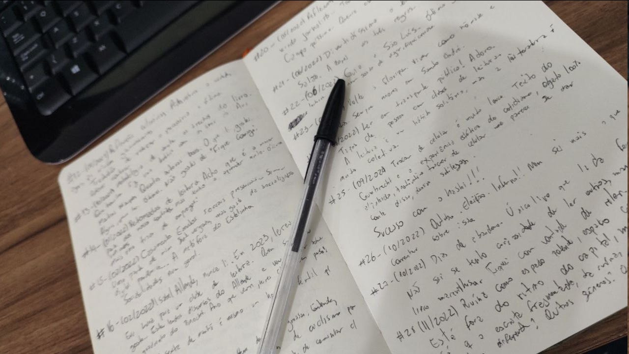 um caderno aberto sobre uma mesa de madeira, com teclado ao fundo. As folhas são brancas, sem linha. A escrita preenche as duas páginas, com uma letra de forma. A tinta é preta, assim como a caneta com tampa preta que repousa em cima do caderno.