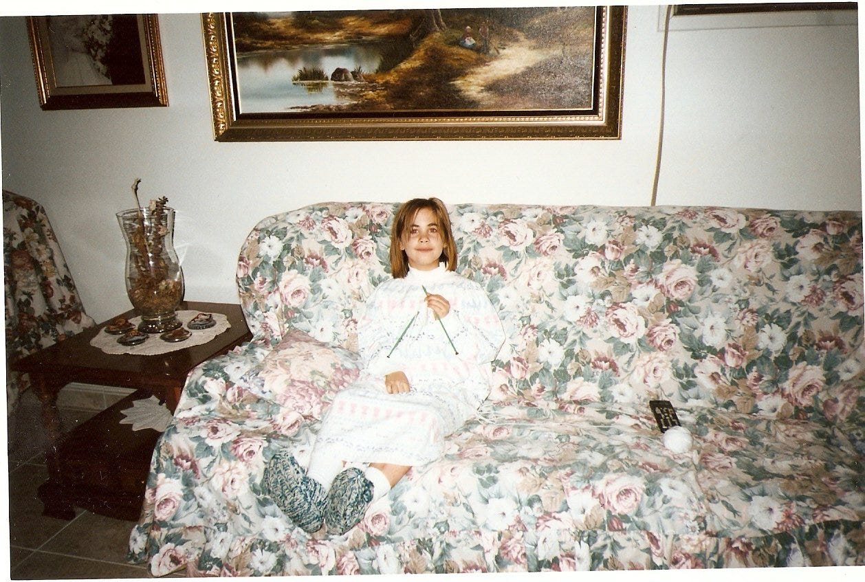 Cassandra as a kid, in a white nightgown and turtle neck, sitting on her grand-parents' couch. The couch is covered with a floral fabric and Cassandra holds two knitting needles, a ball of yarn to her left. The end table beside her has a doily on it.