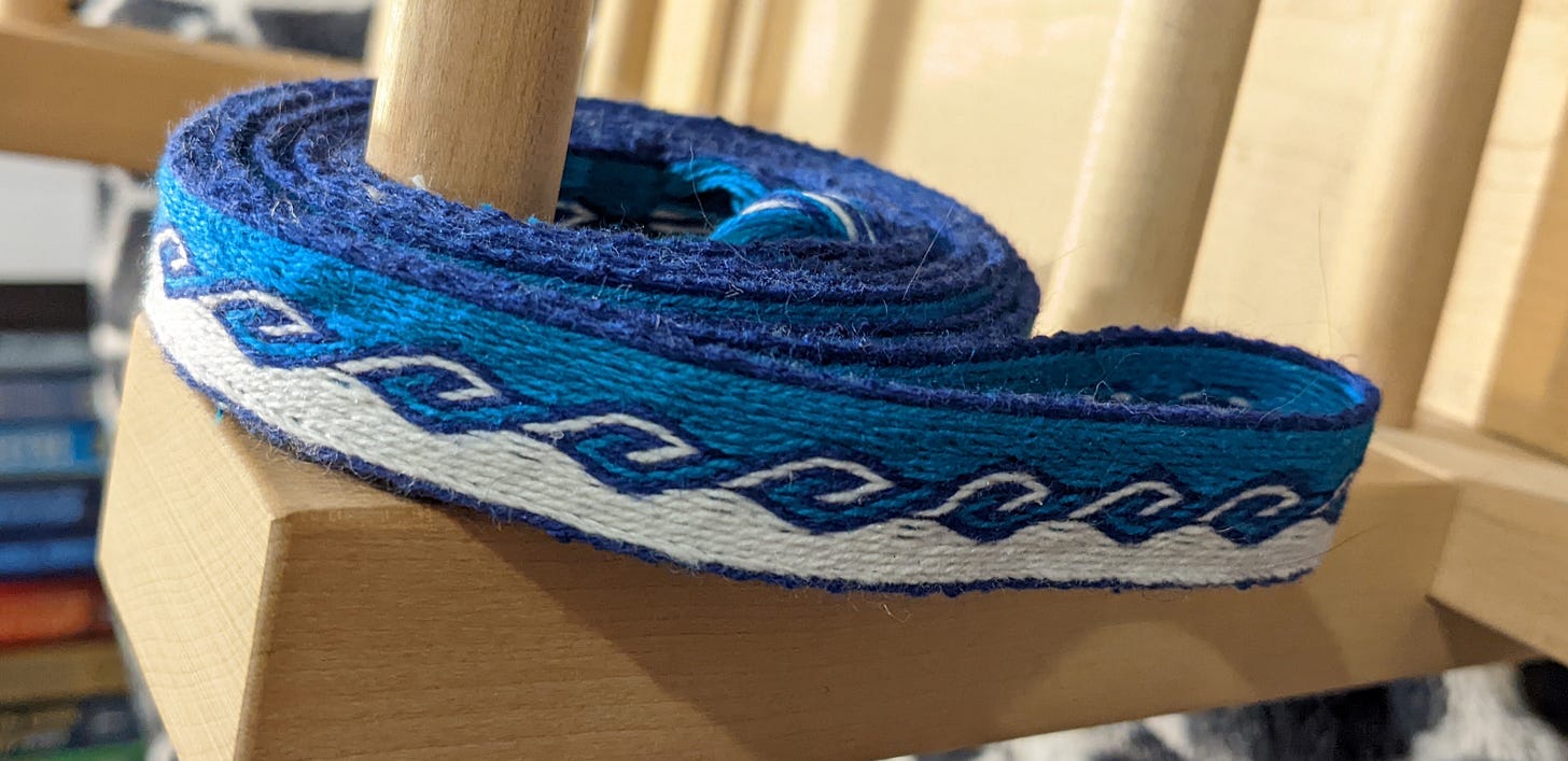 A coil of woven band, an inch wide, in white, turquoise, and dark blue sits on a wooden loom.
