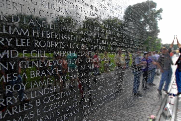 A shiny black granite wall extends from the foreground to the background of the frame with thousands of names etched into it. We can see the reflection of many people in the shininess of the granite.