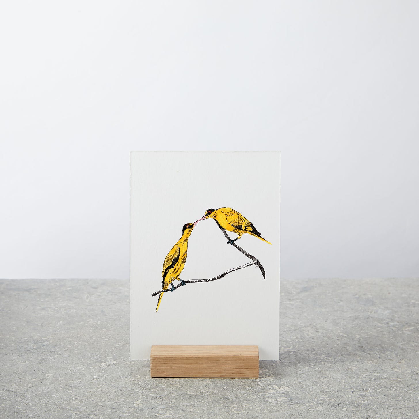 image: photo of a mini art print on a wooden stand. It features my artwork of Commitment, a pair of yellow oriole birds on a single tree branch. Their beaks are touching each other.