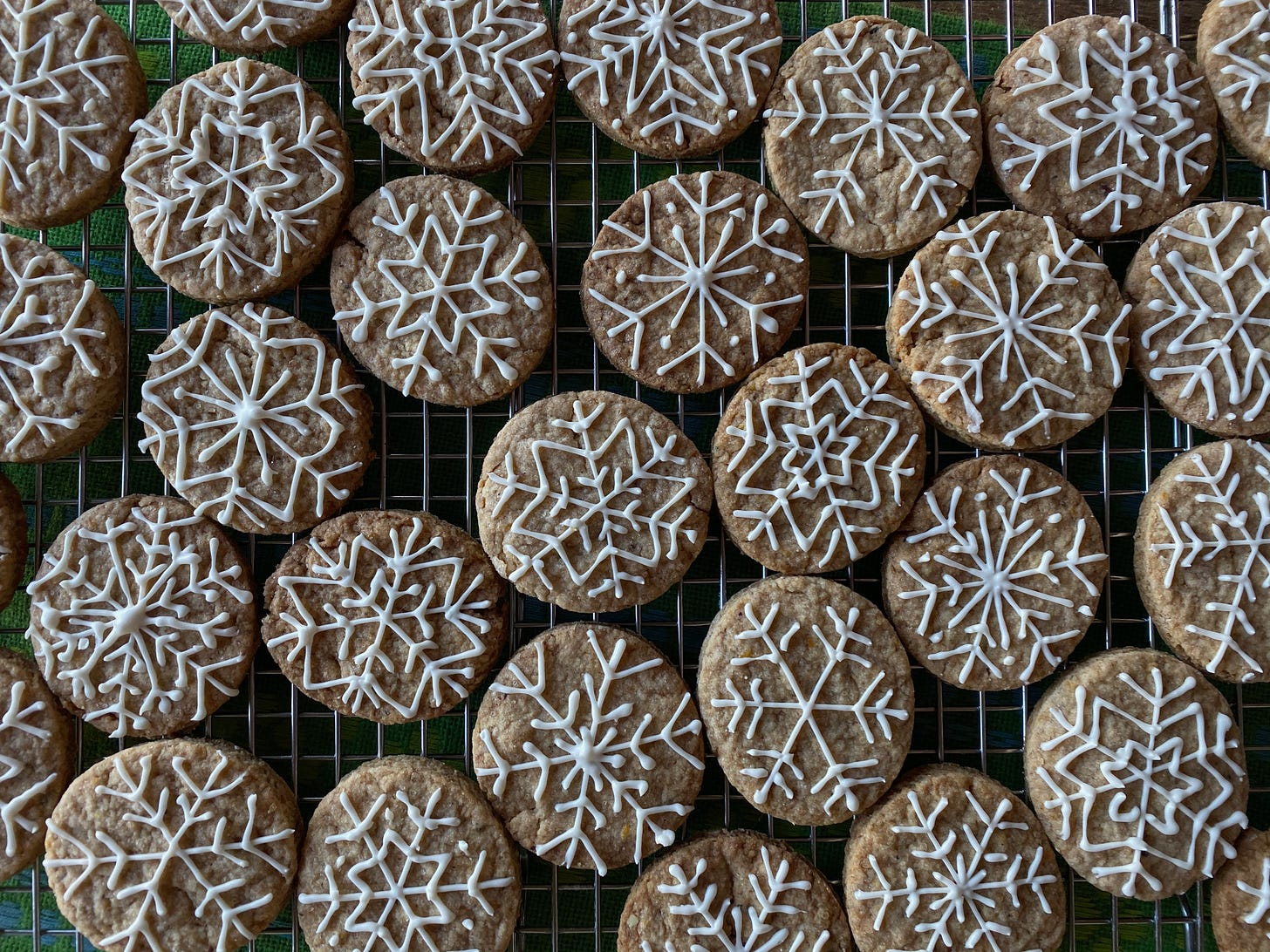 Many round golden brown cookies sit on a metal cooling rack. They are intricately decorated in white icing with different snowflake patterns.