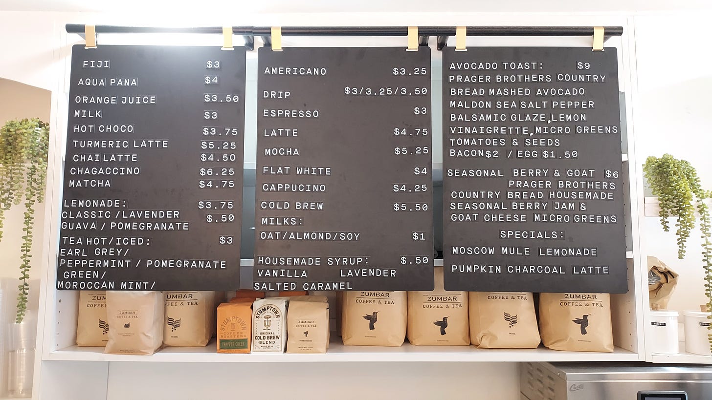 Close up of a three-panel black hanging menu board with the drink and food offerings listed in white block lettering on the left and prices on the right of each panel. A white shelf behind and just below the hanging menuboard is loaded with cardboard brown coffee bags with the Zumbar Coffee & Tean name and logo. To each side of the frame and shelf a hanging fern hangs down.