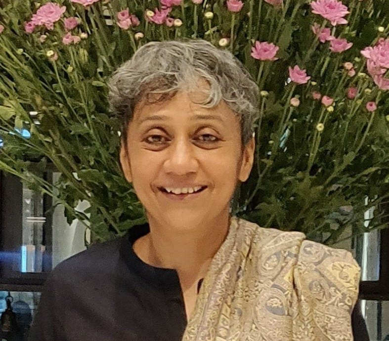 A profile picture of Bhargavi, a brown-skinned woman looking towards the camera. Her mouth is half-open, almost smiling, and she has short hair, mostly grey, tousled slightly. She wears a black top and on one shoulder has draped a shawl with intricate golden patterns. In the background, almost sprouting from her back, are the green stems of pink flowers. 