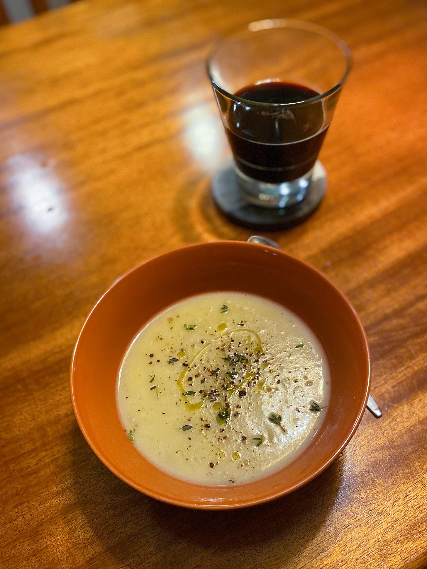 An orange bowl of potato soup, drizzled with oil and flecked on top with cracked pepper and fresh thyme leaves. On a coaster behind the bowl is a glass of stout.