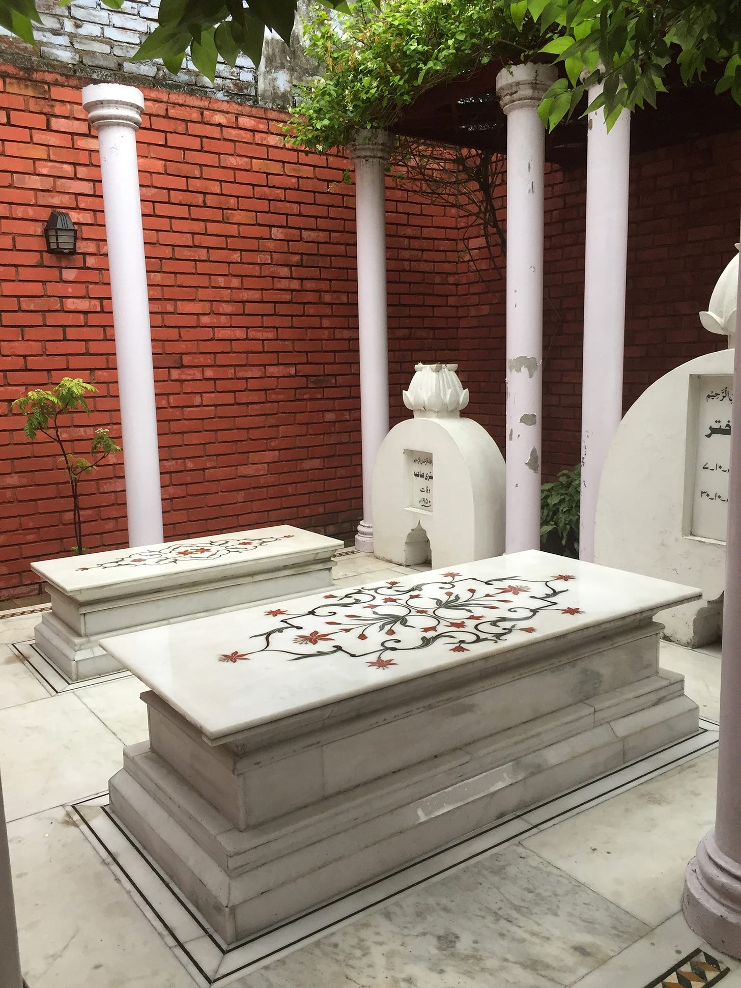 A picture of two marble pietra dura tombs surrounded by red brick walls and lush green trees.