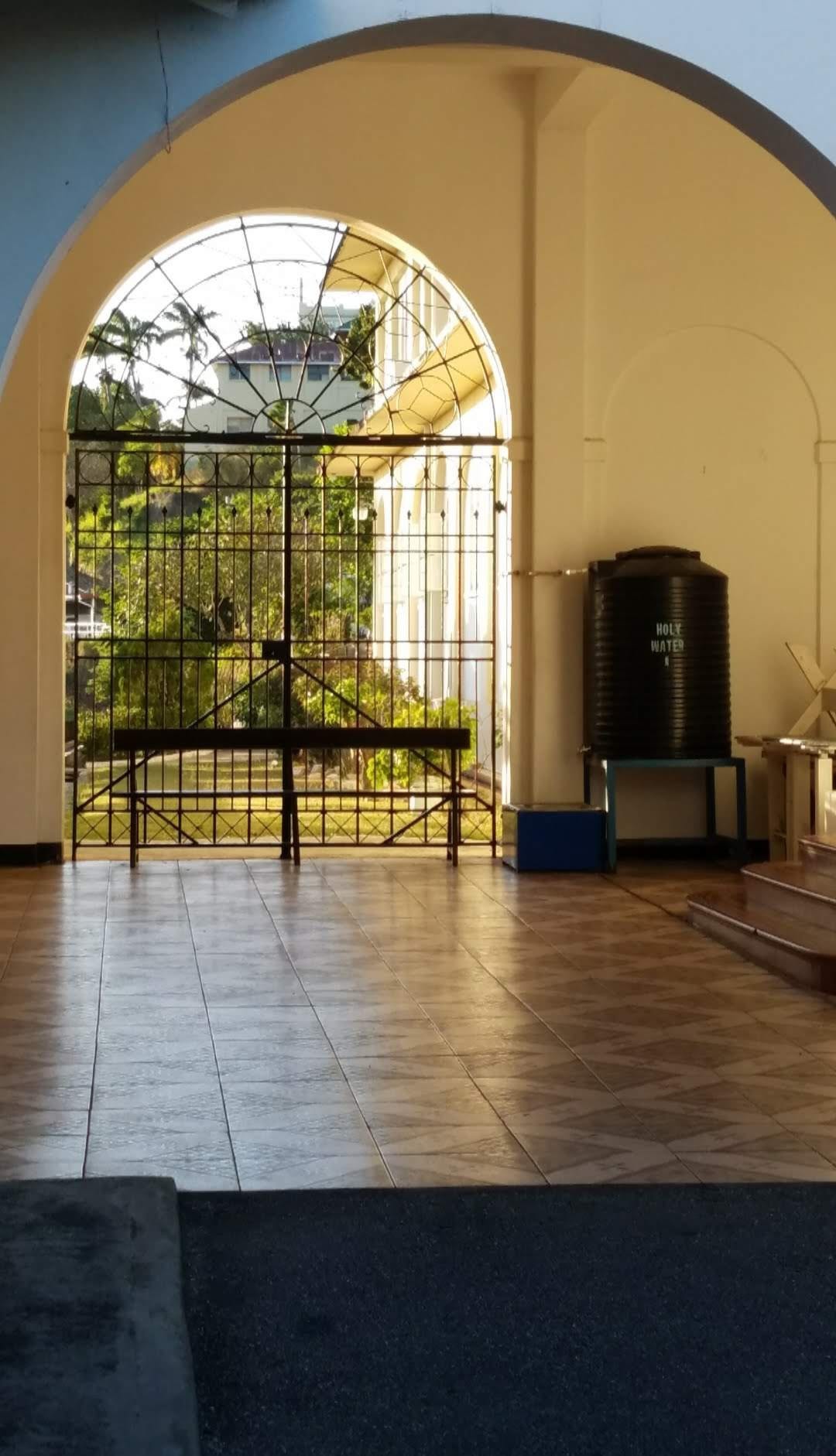 A black iron gate with a curved half circle on top is visible through a white archway. The floors are brown tile. Sunlight reflects on them. Trees are visible through the gate.