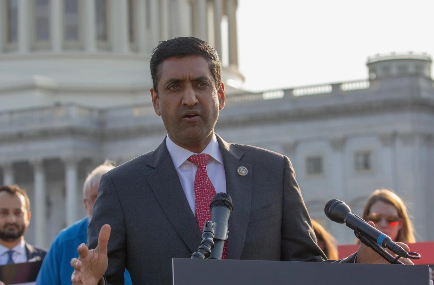Congressman Ro Khanna stands at a podium in front of microphones at the capitol in Washington. He is wearing a dark suit and a red tie. Behind him is a crowd of people and the capitol building.