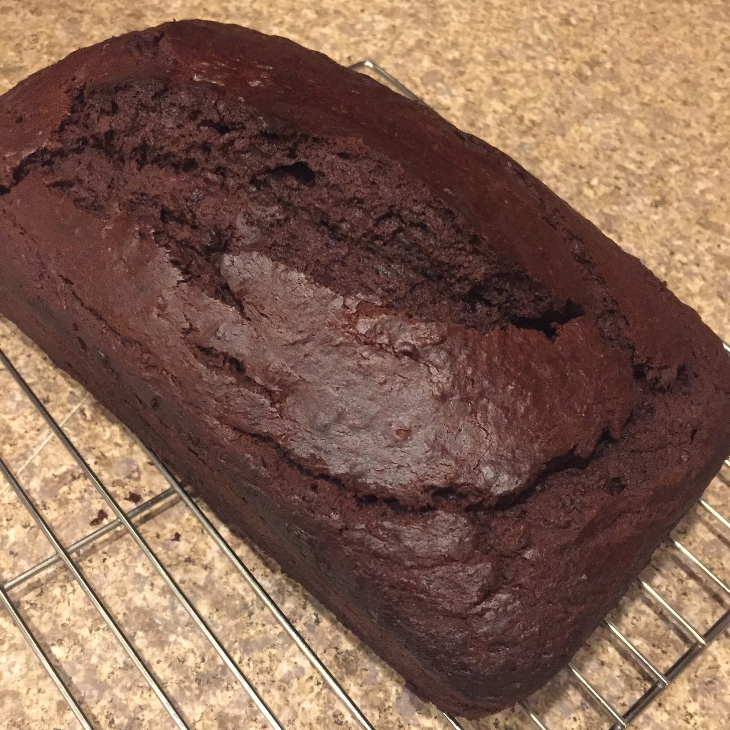 on a cooling rack, a dark brown loaf of chocolate banana bread, cracked open across the top.
