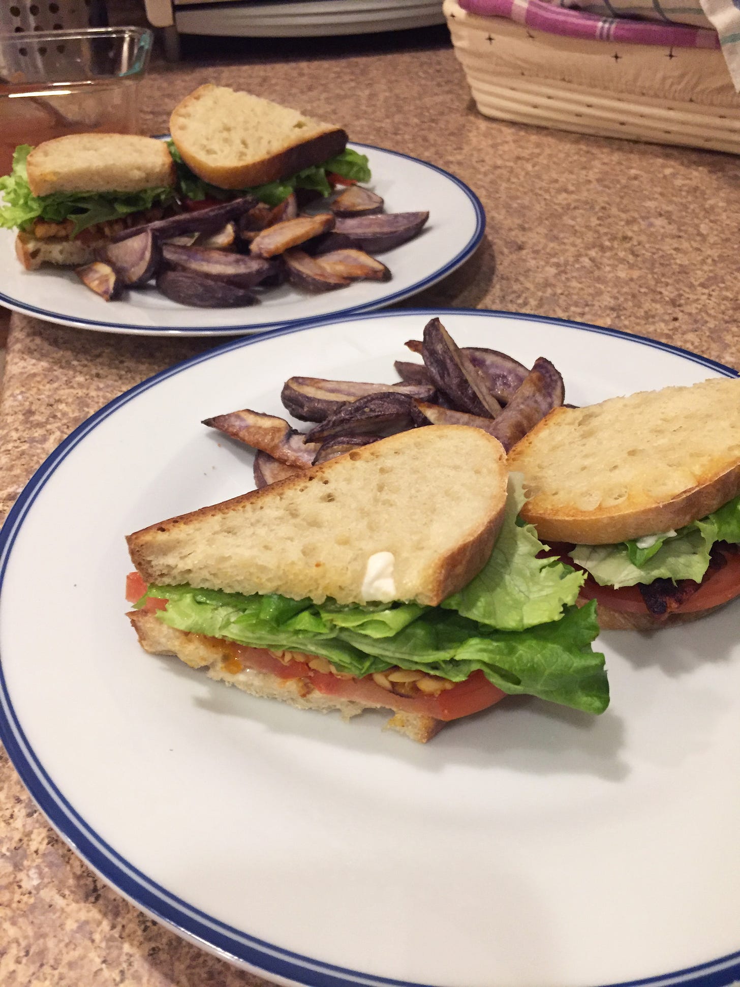Two dinner plates, each with a sandwich cut in half next to a pile of purple potato fries. The open side of the sandwich shows tomato, tempeh, and lettuce.