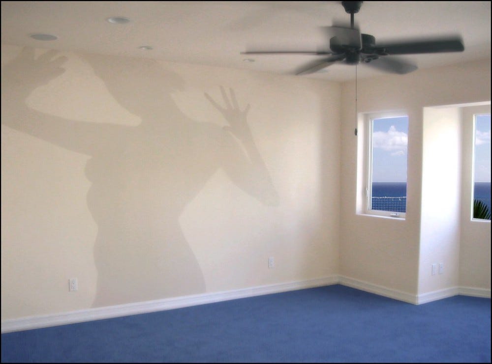 Empty room with blue carpet and small windows overlooking water. A blurry ceiling fan appears to be in motion. The shadow of a woman holding her hands up is cast on the far wall.