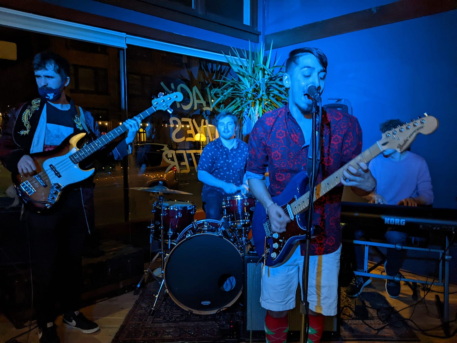 A four piece band with guitar, keyboard, bass, and drums, playing in front of a window