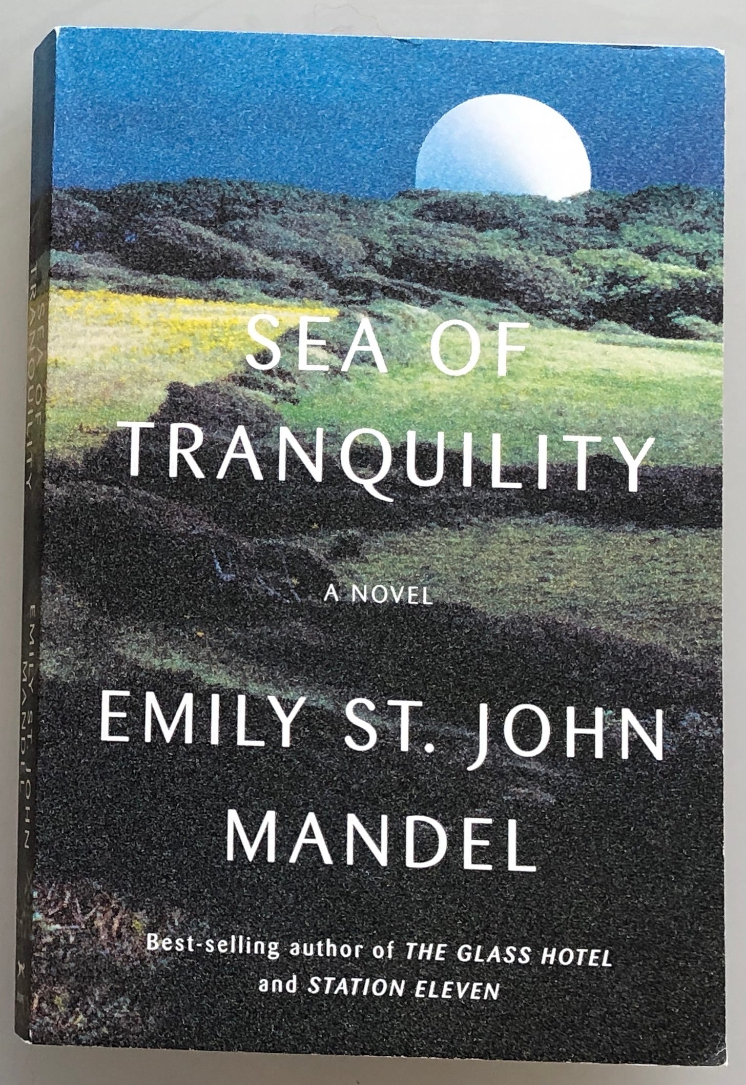 A green and yellow hillside and blue sky with moon rising compose this book cover. Notes that St. John Mandel is the best-selling author of The Glass Hotel and Station 11.