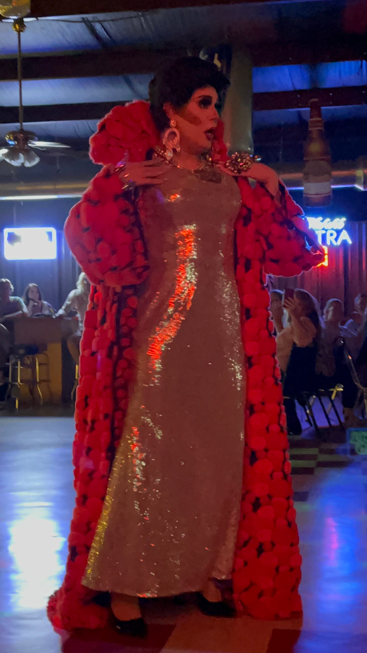 A drag queen with black hair and a fluffy robe performs in front of a crowd in the dimly lit Good Times Bar.