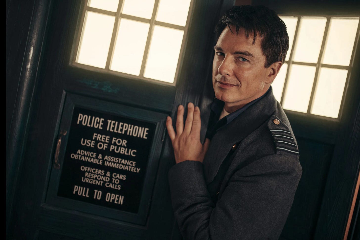 This image shows Captain Jack Harkness (played by John Barrowman), with a devil-may-care smile on his face, standing half-in and half-out of the TARDIS's front door. This image shows Captain Jack Harkness (played by John Barrowman), with a devil-may-care smile on his face, standing half-in and half-out of the TARDIS's front door.