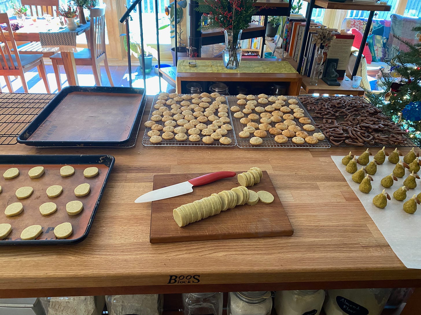 A sunlit kitchen island full of trays of cookies. There are small marzipan pears, a tray of cocoa alphabet cookies, two cooling racks full of small almonds cookies, an empty tray, and a log of sliced unbaked cookies with a knife on a cutting board.