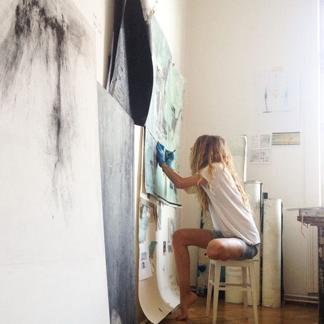 Side portrait of a woman with blonde curly hair with gloves on, sitting on a stool, drawing with charcoal in an artist studio