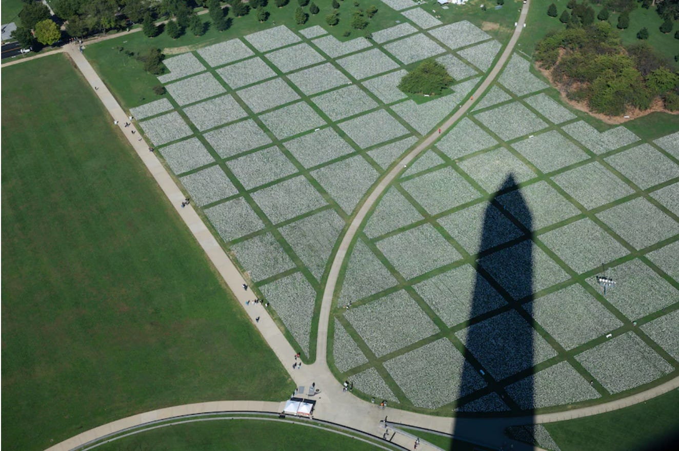 An aerial view that shows a large white grid with sections of green grass in between. The tip of the Washington Monument obelisk can be seen in shadow looming over the ground.