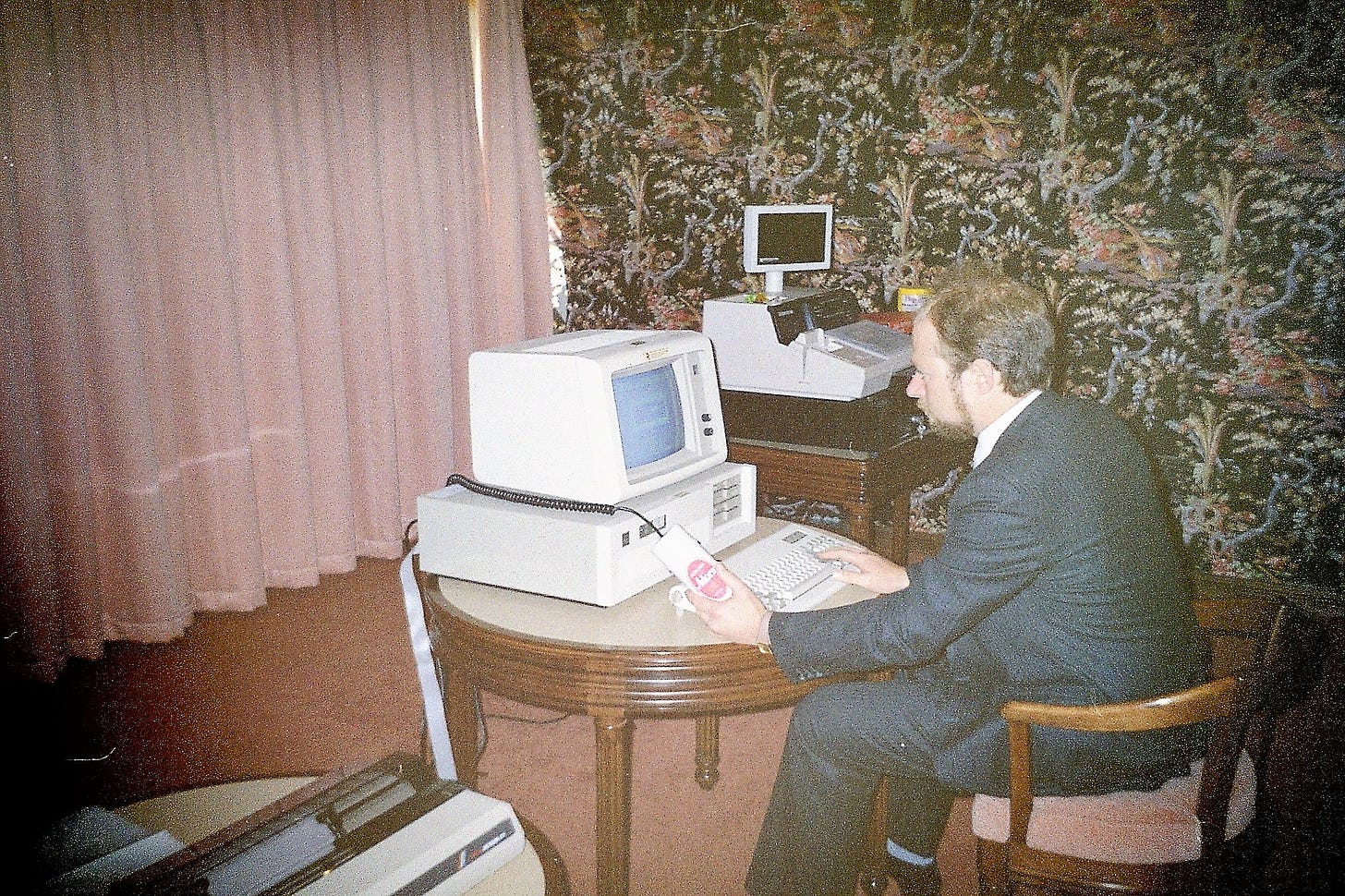 Luigi Cappel, in a suit, typing on an old computer surrounded by pink and flowery curtains.
