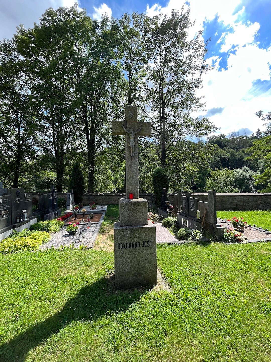 Cemetery with cement cross in the middle, tall trees around the fence, blue sky
