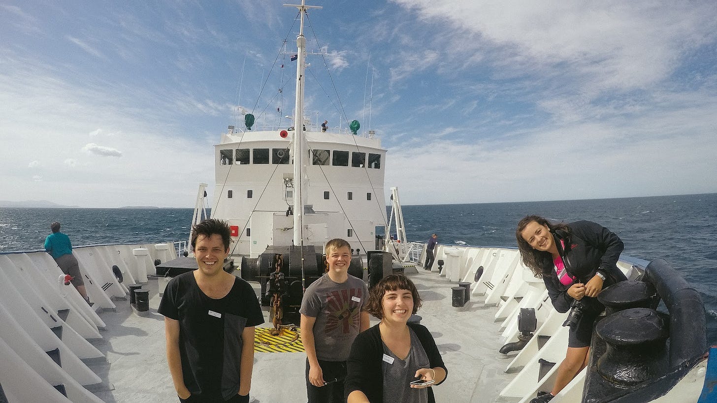 A photo taken on a fish-eye GoPro, with the wide-angle capturing the width of the ship, with four young people standing on the bow, looking at the camera - the one holding the handle of the GoPro is Louise. Behind them you can see the horizon, the dark blue ocean, and a blue sky with white clouds.
