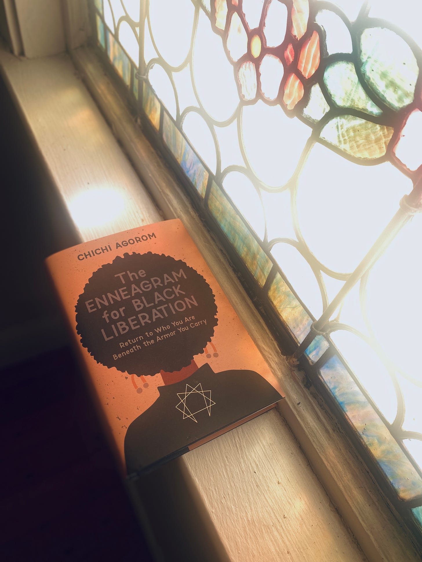 A copy of the Enneagram for black liberation. Sitting on a windowsill, the window I stain glass with light pouring through. Some of the pink Stainglass matches the pink background the cover of the book, which has a graphic of a woman's Afro black shirt gram symbol on shirt in the title of the book in the Afro.