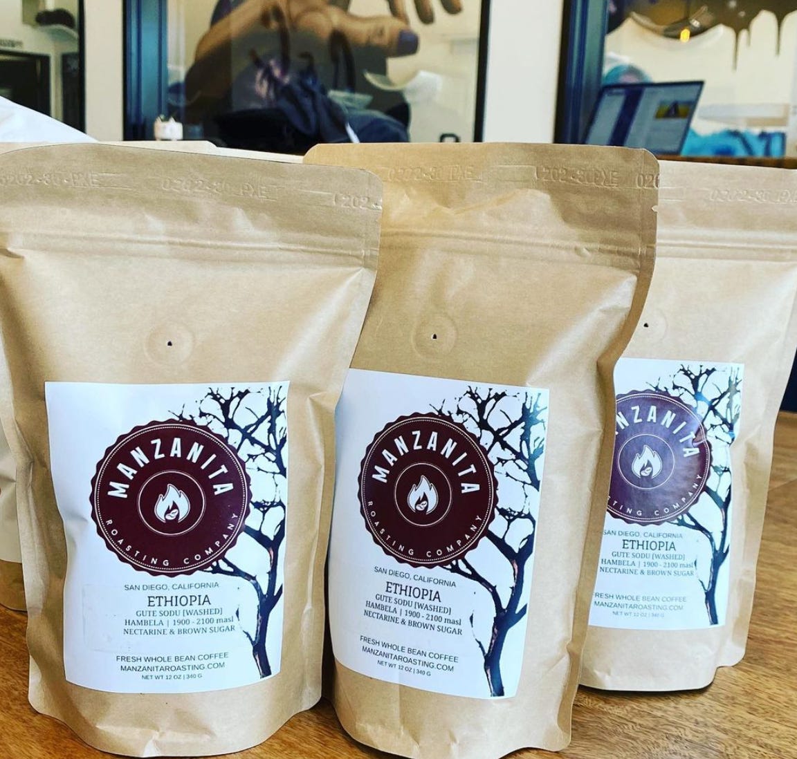 Three brown, vacuum sealed bags of coffee with a white square label and the Manzanita Coffee Roasting label (maroon) on it side by side by side. Close up.