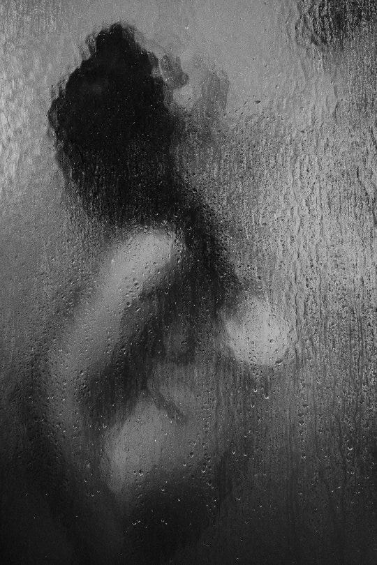 Descrição da imagem: em preto e branco. O corpo de uma mulher de perfil num box de chuveiro, onde ela está de lado, olhando pra cima. Cabelos negros e uma fonte de luz vinda de cima, mais para a direita da foto.