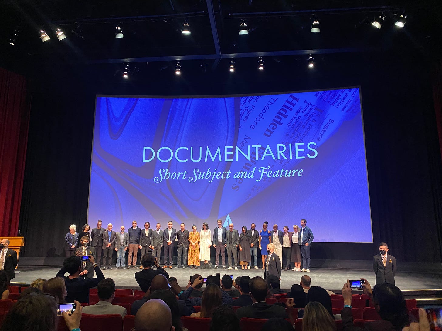 Inside a cinema, people line up on stage to have their photo taken. The words "Documentaries Short Subject and Feature" is on the background.