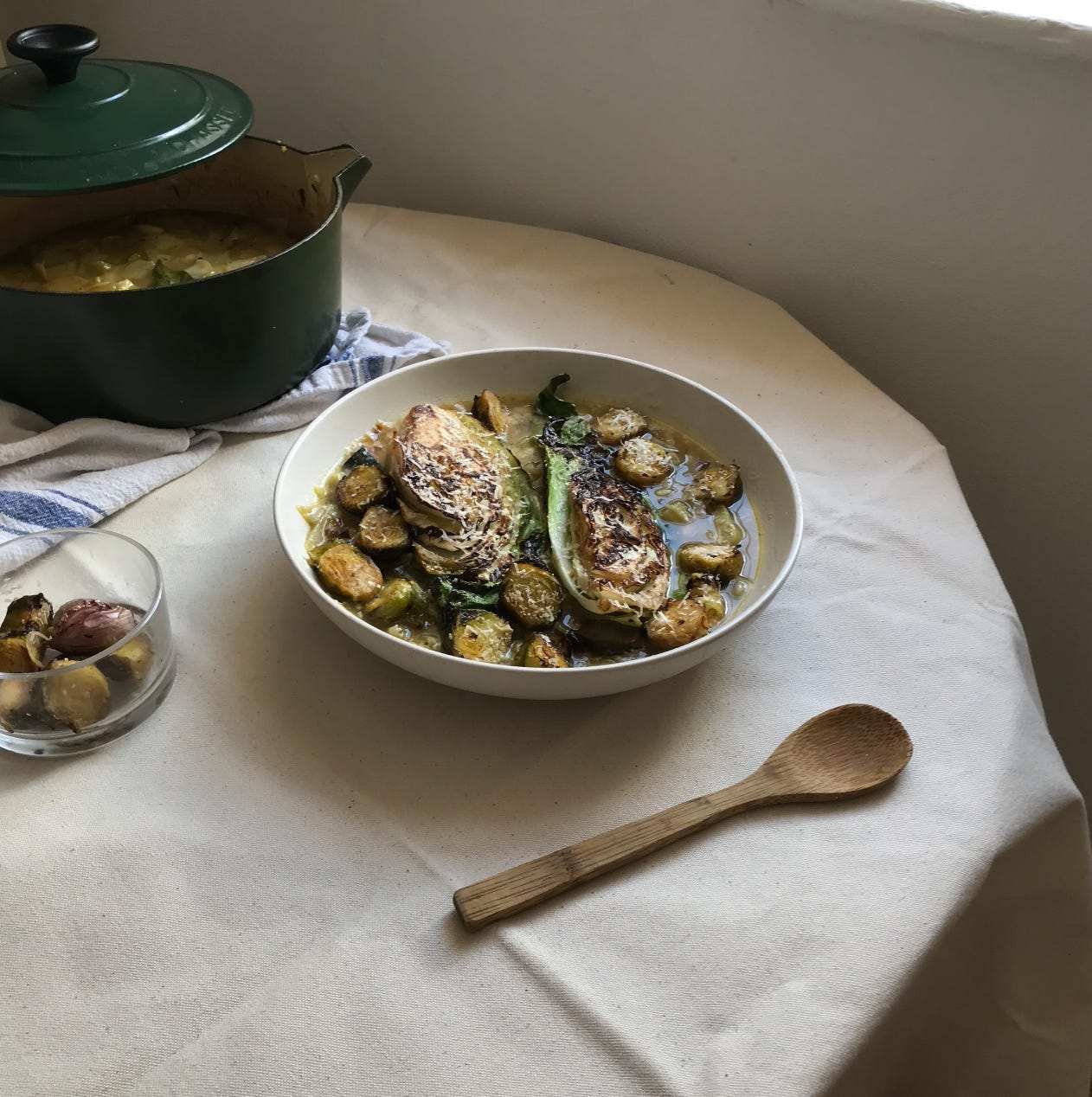 A photo of a large shallow dish on a table. Beside it, a bottle green casserole dish, lid half on, a wooden spoon, and a glass ramekin with potatoes in. In the bowl, a liquidy mix, yellow and green, are coins of courgette, and charred halves of hispi cabbage. 