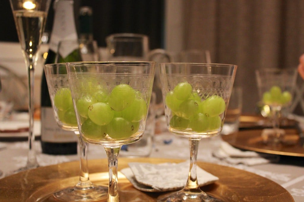 A table with a white tablecloth, a bottle of champagne, a champagne flute, and several wine glasses with 12 grapes in each of them, arranged on gold plates.