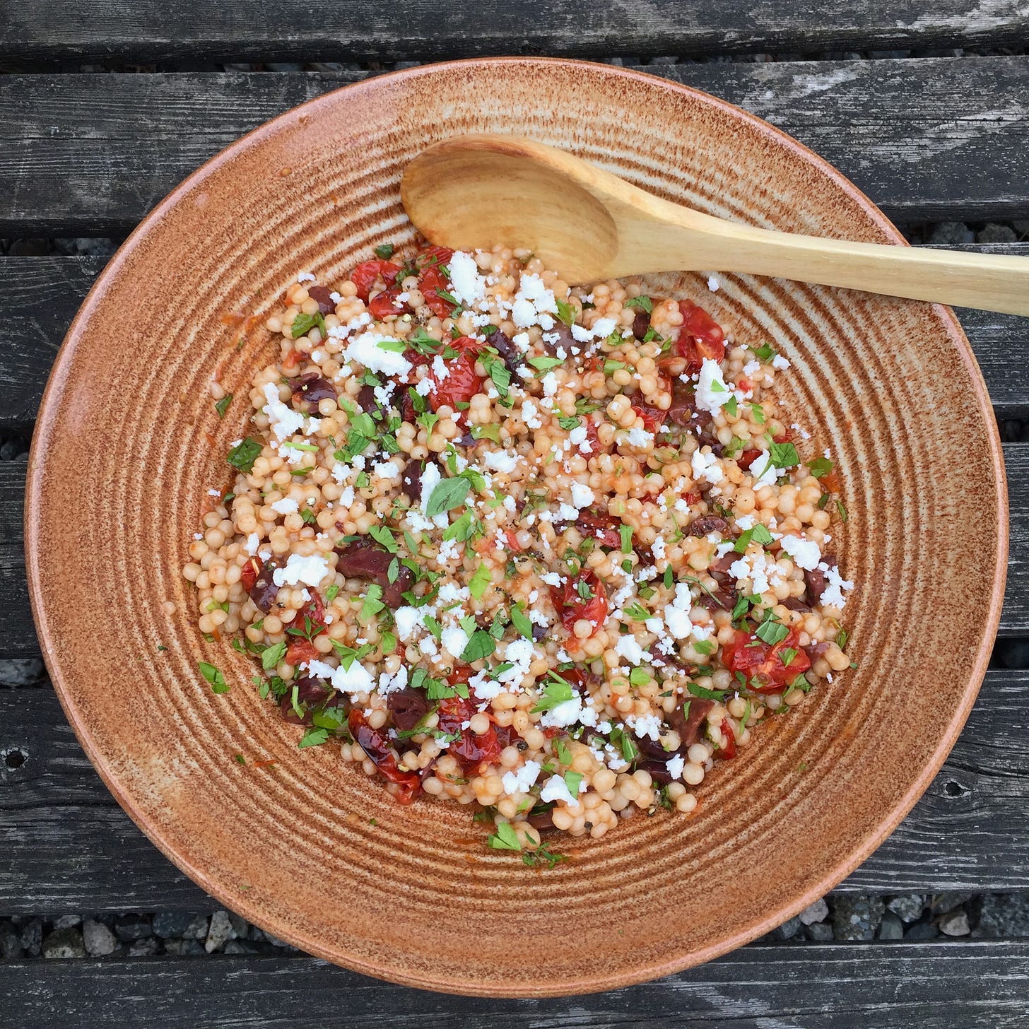 A large stoneware bowl of pearl cousous tossed with kalamata olives, pieces of grape tomato, and herbs. Crumbles of feta and more herbs are sprinkled over the top, and a wooden spoon rests at the top edge of the bowl.