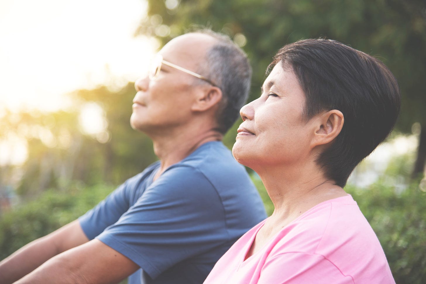 Elderly Asian couple with eyes closed, sitting in a park, content in the sunrise, breathing.