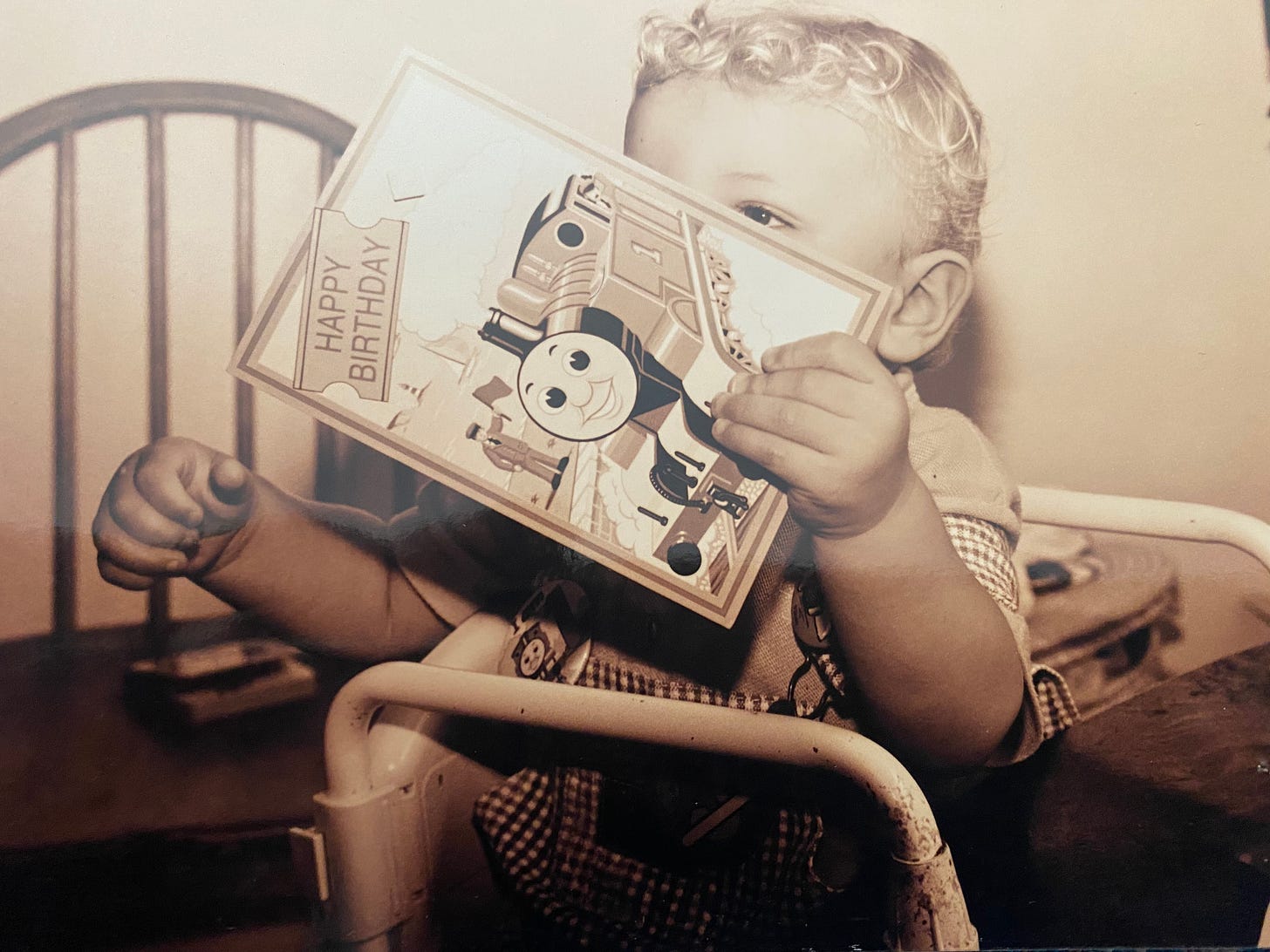 A blond curl-haired baby holds a Thomas The Tank 1-year birthday card covering his face and one eye.
