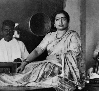 A picture of Gauhar Jaan dressed in a saree sitting on the floor next to an accompanist and a phonograph.