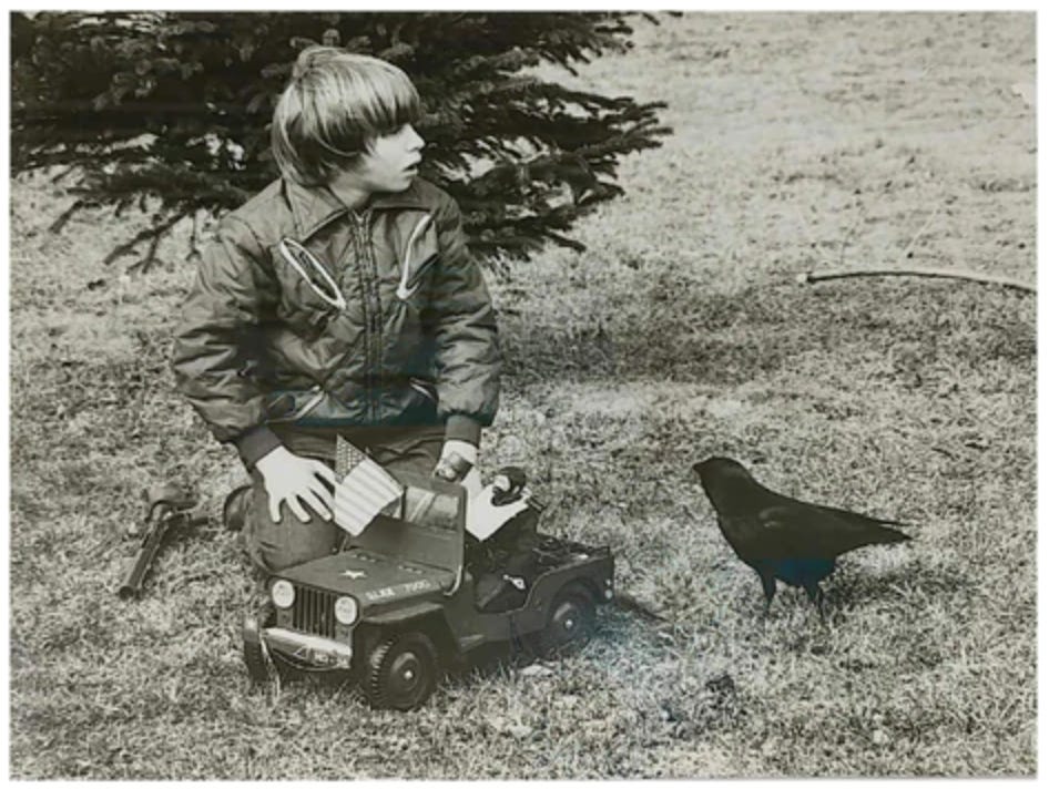 A black and white photo of a boy on his knees in the lawn with a GI Joe toy jeep and Joe. A crow looks up at him from near his knee.