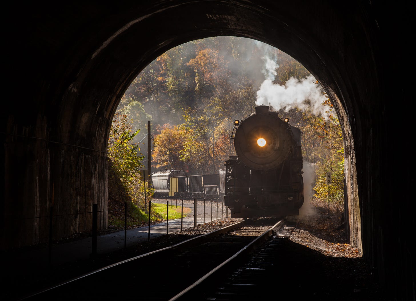 Old train entering a tunnel with a single headlight on and smoke billowing from the engine