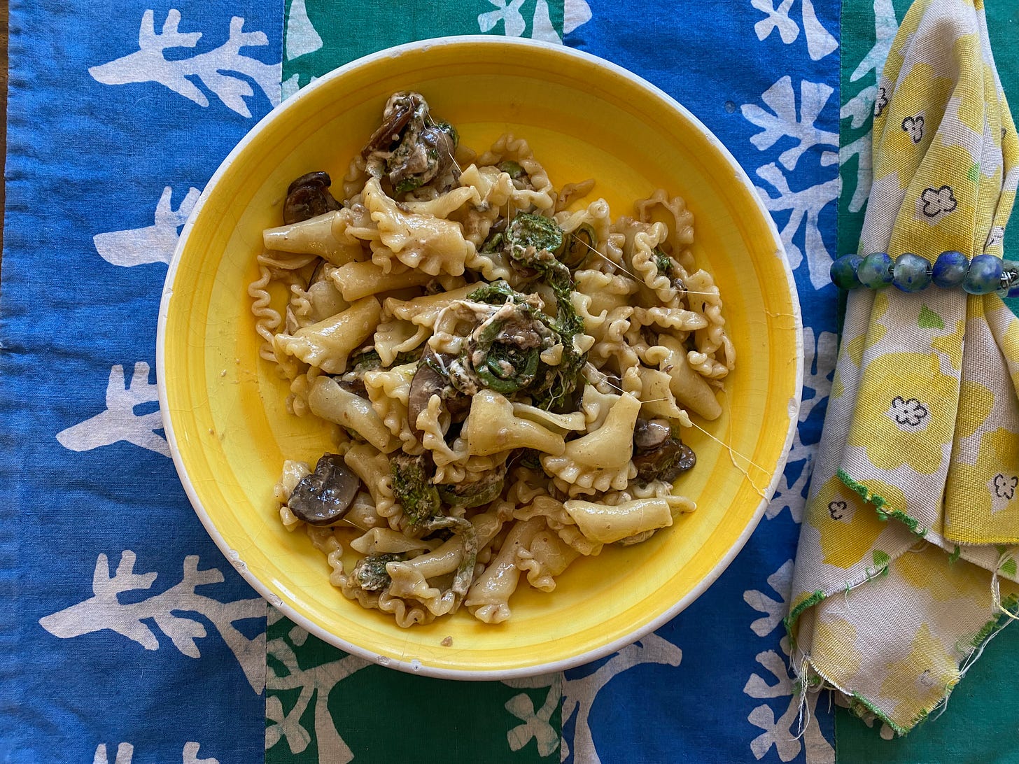 A shallow yellow bowl of cheesy pasta, mushrooms, and fiddlehead ferns sits on a blue and green placemat next to a yellow cloth napkin.